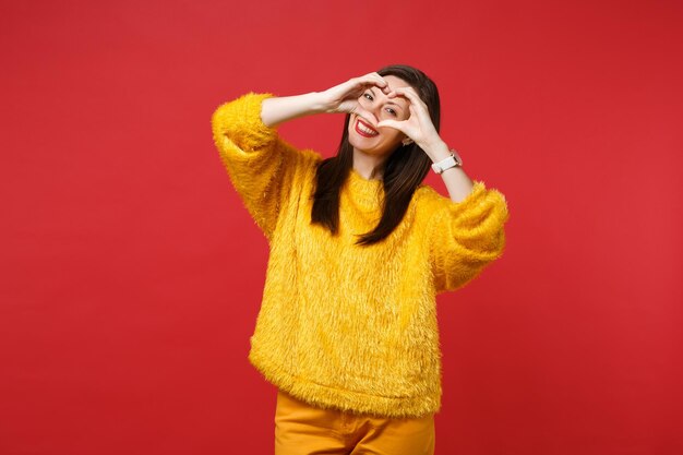 Portrait of pleasant young woman in yellow fur sweater showing shape heart with hands isolated on bright red wall background in studio. People sincere emotions, lifestyle concept. Mock up copy space.