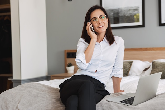 Portrait of pleased adult businesswoman in formal suit talking on smartphone and using laptop while sitting on bed in apartment