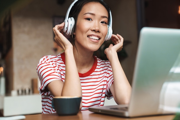 Portrait of pleased asian woman listening to music with headphones while using laptop computer in cafe indoors