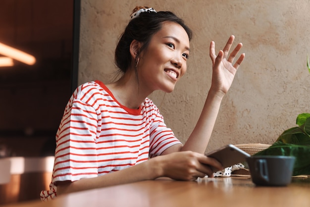 Portrait of pleased asian woman waving hand and holding cellphone while sitting in cafe indoors