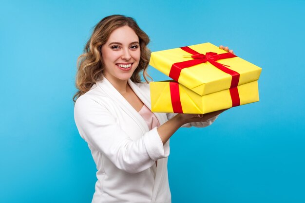 Portrait of pleased charming woman with wavy hair in white jacket unpacking big present and cute smiling at camera satisfied with great holiday gift indoor studio shot isolated on blue background