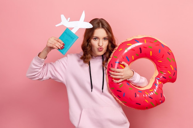 Portrait of pleased curly haired teenage girl in hoodie with pout lips showing donut rubber ring passport and paper airplane happy with future trip Indoor studio shot isolated on pink background