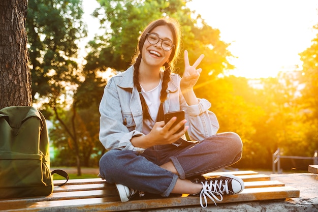 Portrait of a pleased cute young student girl wearing eyeglasses sitting on bench outdoors in nature park with beautiful sunlight using mobile phone showing peace sign