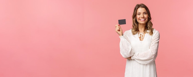 Foto il ritratto di una bella femmina europea bionda in abito bianco mostra la carta di credito con l'espressione soddisfatta sorridente fotocamera consiglia i servizi bancari utilizzare il pagamento online sfondo rosa