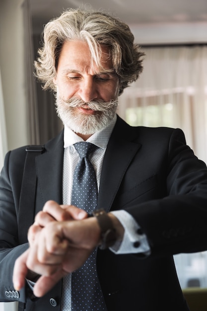 Portrait of a pleased handsome grey-haired senior businessman indoors at home dressed in formal clothes looking at watch clock.