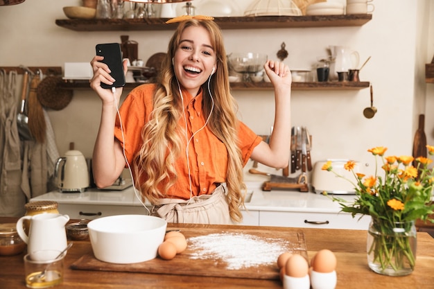 Portrait of a pleased happy young blonde girl chef cooking at the kitchen listening music with earphones using mobile phone.