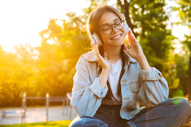 Portrait of a pleased happy young teenage girl student sitting outdoors in beautiful green park listening music with headphones