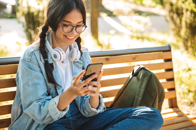 Portrait of a pleased positive cheerful cute young student girl wearing eyeglasses sitting on bench outdoors in nature park using mobile phone.