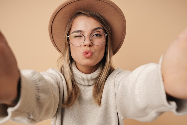 Portrait of pleased young woman in hat winking and taking selfie photo 