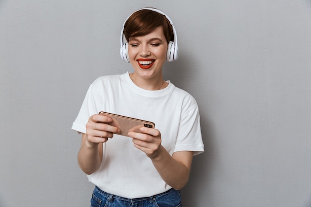 Portrait of pleased young woman wearing headphones playing video games on cellphone isolated over gray wall