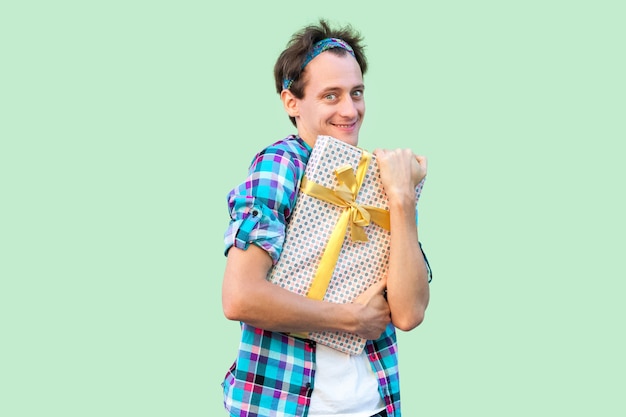 Portrait of pleasure young hipster man in white t-shirt and checkered shirt standing and hugging present with yellow bow, sly smile, looking at camera. Indoor, isolated, studio shot,green background