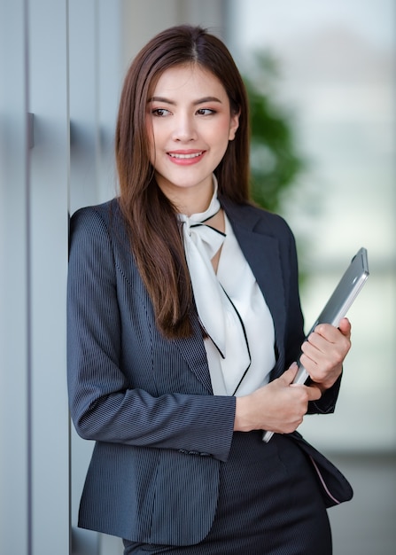 Portrait of positive asian female entrepreneur or secretary in\
formal clothes standing with laptop netbook computer in office hall\
and looking at far point in relax pose.