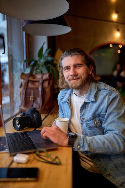 Portrait of positive bearded guy looking at camera while working on laptop in cafe