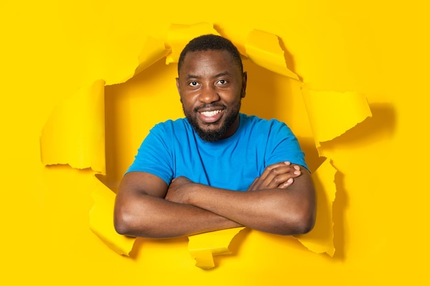 Portrait of positive black man posing with folded arms in torn\
paper hole looking and smiling at camera