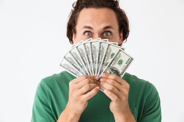 Portrait of positive brunette man wearing basic t-shirt covering his face with cash money isolated over white wall