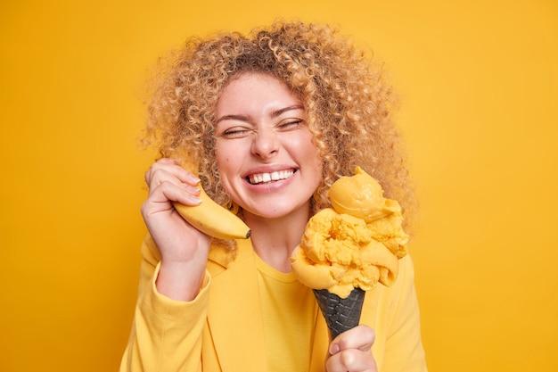 Portrait of positive Caucasian woman smiles broadly shows white even teeth being in good mood as eats favorite dessert holds banana near ear closes eyes from pleasure isolated over yellow wall