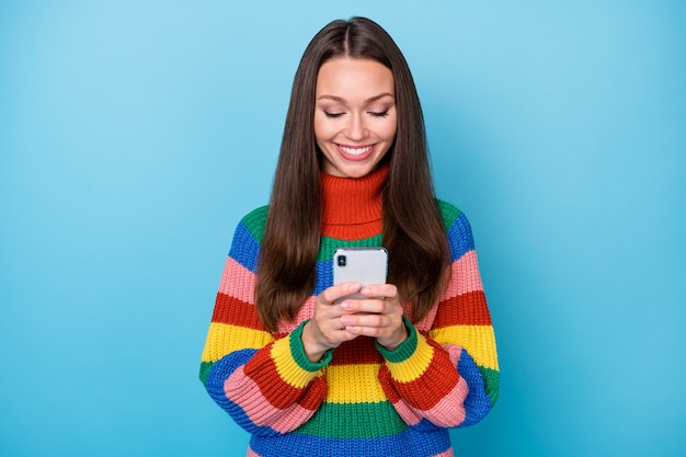 스마트폰을 사용하는 긍정적인 쾌활한 소녀 블로거의 초상화는 소셜 미디어 뉴스를 구독하고 파란색 배경에 격리된 무지개 스타일의 스웨터를 입습니다.