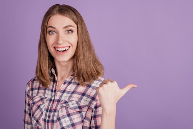 Portrait of positive cheerful girl indicate thumb empty space present adverts promo isolated over violet color background