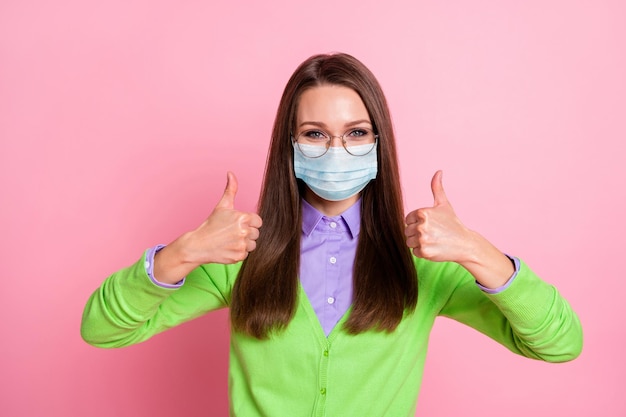 Il ritratto di una ragazza allegra positiva mostra il segno del pollice in su indossa una maschera medica isolata su uno sfondo di colore pastello