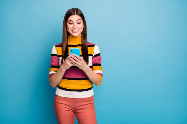 ポジティブで陽気な女の子の肖像画はスマートフォンを使用しますソーシャルネットワーク情報を読みますコメントを共有しますフィードバックは青い色の壁に隔離されたスタイリッシュな赤いジャンパーを着用します