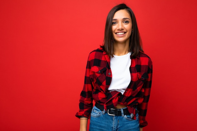 Portrait of positive cheerful smiling young brunette woman in casual white t-shirt, stylish red check shirt isolated on red background with copy space.