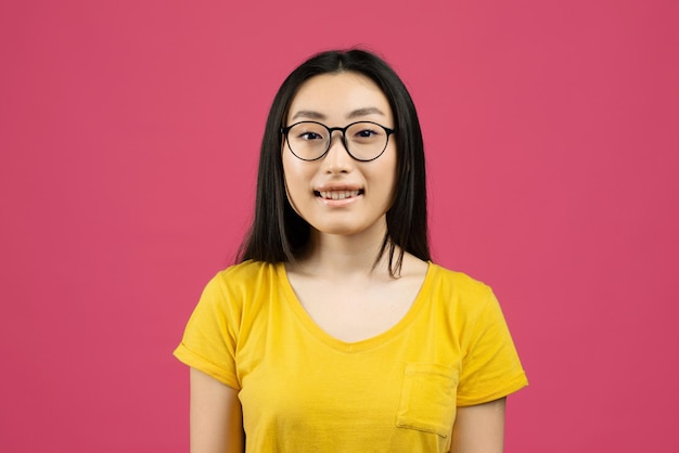 Портрет позитивной китаянки в очках и смотрящей в камеру, позирующей на розовом студийном фоне