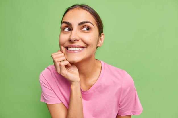 Portrait of positive cute brunette young woman keeps hand under chin smiles broadly imagines something good or pleasant wears pink t shirt isolated over green wall