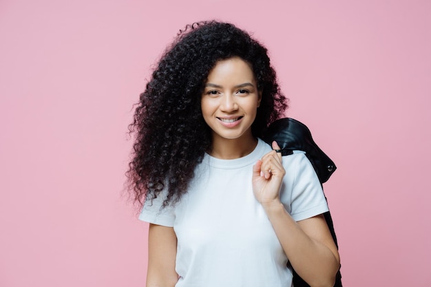 Portrait of positive ethnic woman wears white t shirt carries jacket on shoulder has cheerful expression ready for picnic enjoys day off poses indoor against rosy background People lifestyle