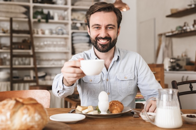 Portrait of positive european man sitting at table and drinking coffee while having breakfast in kitchen at home