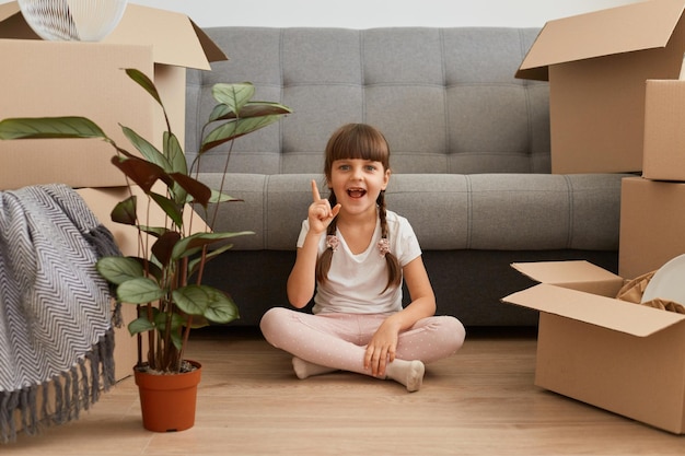 Portrait of positive excited little girl wearing casual style attire sitting on a floor with raised finger, pointing up, having idea of new room decor, expressing positive emotions.