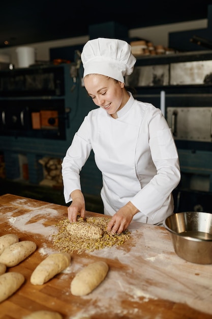 Portrait of positive female baker shaping bread bagels, dipping dough with seeds and oatmeal flakes. Bakery house concept