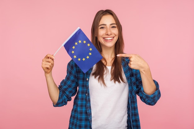 Ritratto di donna amichevole positiva in camicia a scacchi che sorride ampiamente e indica la bandiera dell'unione europea simbolo dell'europa ue associazione comunità indoor studio girato isolato su sfondo rosa