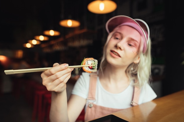 portrait of positive girl in cap and pink sundress eating sushi with chopsticks in Asian cafe