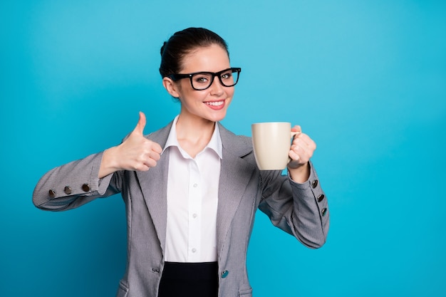 Il ritratto della tazza positiva della tenuta della ragazza mostra il pollice sul segno isolato sopra il fondo di colore blu