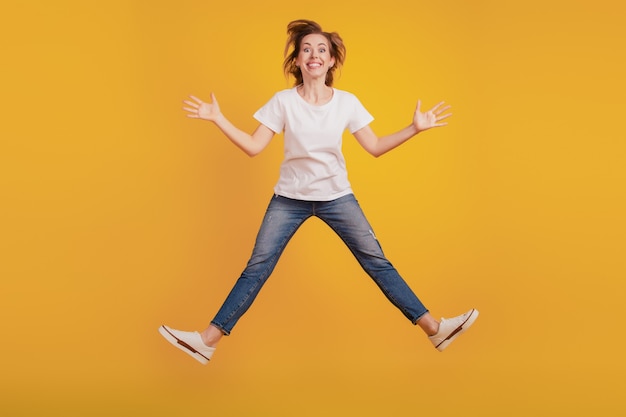 점프하는 긍정적인 소녀의 초상화는 노란색 배경에 손을 들고 이빨 미소를 짓습니다.