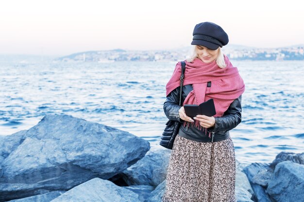 Portrait of positive and happy mature woman with smartphone at the beach. Autumn, winter season on the sea.