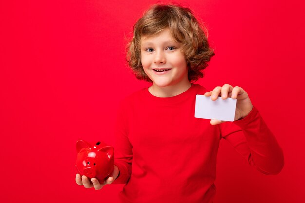 Portrait of positive happy smiling little boy with curly blond hair with sincere emotions wearing casual red pullover isolated over red background with copy space, holds red piggy box for coins and cr