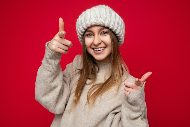 Portrait of positive happy smiling young beautiful dark blonde lady with sincere emotions wearing beige jersey and knitted hat isolated over red background with copy space and pointing at camera