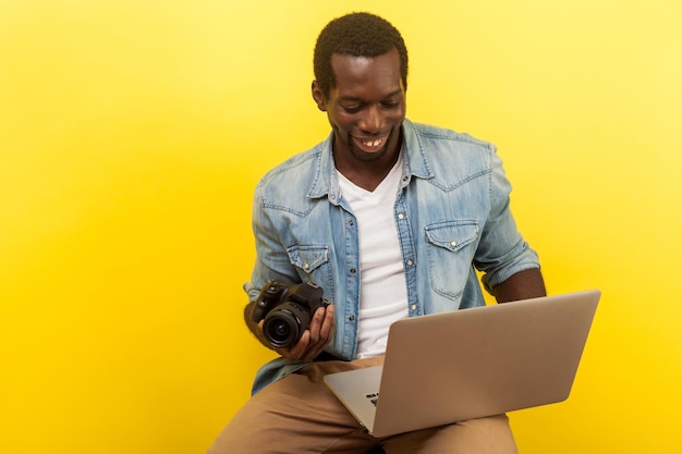 Ritratto di fotografo maschio positivo in camicia di jeans con fotocamera digitale dslr e laptop per scattare foto utilizzando l'app di fotoritocco su computer studio girato isolato su sfondo giallo