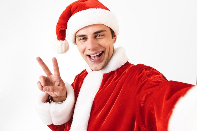 Portrait of positive man 30s in santa claus costume and red hat laughing while taking selfie photo