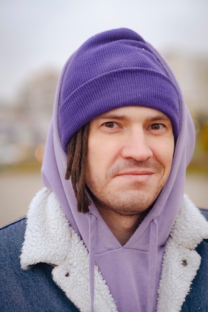 Portrait of positive man with dreadlocks in hat warm clothes on city street Happy male looking at camera smiling in cloudy snowy weather