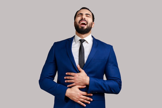 Portrait of positive optimistic bearded man laughing loud hearing really funny thing keeps hands on belly wearing official style suit Indoor studio shot isolated on gray background