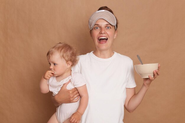 Portrait of positive optimistic satisfied woman wearing white T shirt standing with her little daughter in hands isolated over brown background feeding her child with puree