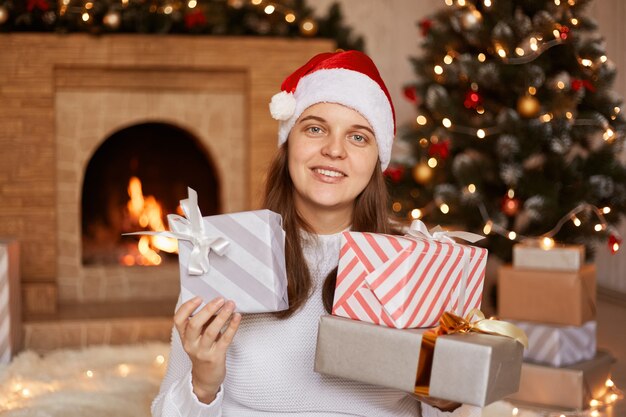 Portrait of positive optimistic woman wearing white sweater and santa claus hat, holding new year presents, being in festive room with fireplace and Christmas tree.