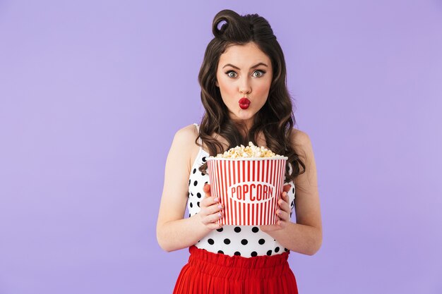 Portrait of positive pin-up woman in retro polka dot dress holding bucket with popcorn while watching movie isolated over violet wall