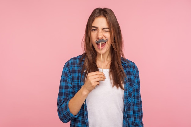 Portrait of positive playful girl in checkered shirt holding fake curled mustache on stick and winking to camera, having fun, wearing masquerade accessory. studio shot isolated on pink background