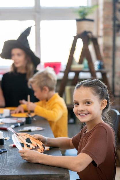 Portrait of positive pretty girl sitting at table against teacher and classmate and making halloween pumpkin craft at art class