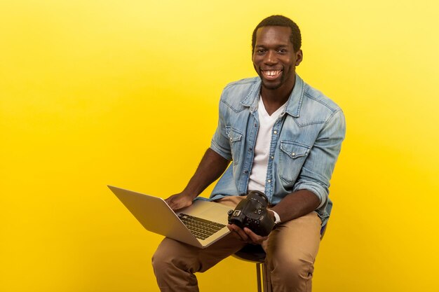 Portrait of positive professional photographer with attractive toothy smile in denim shirt holding digital dslr camera and laptop going to make photo or video indoor studio shot on yellow background