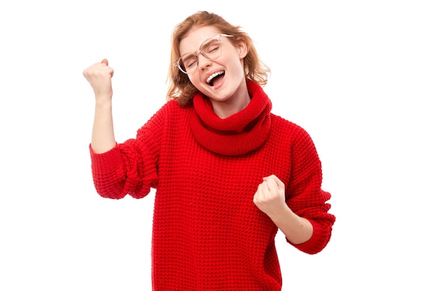 빨간 크리스마스 스웨터를 입은 긍정적인 빨간 머리 소녀의 초상화는 감정적으로 기뻐하고 흰색 배경 광고 배너에 격리되어 행복하다고 느낍니다