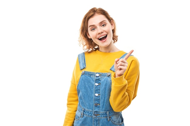 Portrait of positive redhead young woman in casual jeans smiling pointing with finger at empty copy space for text or product isolated on white background advertising banner